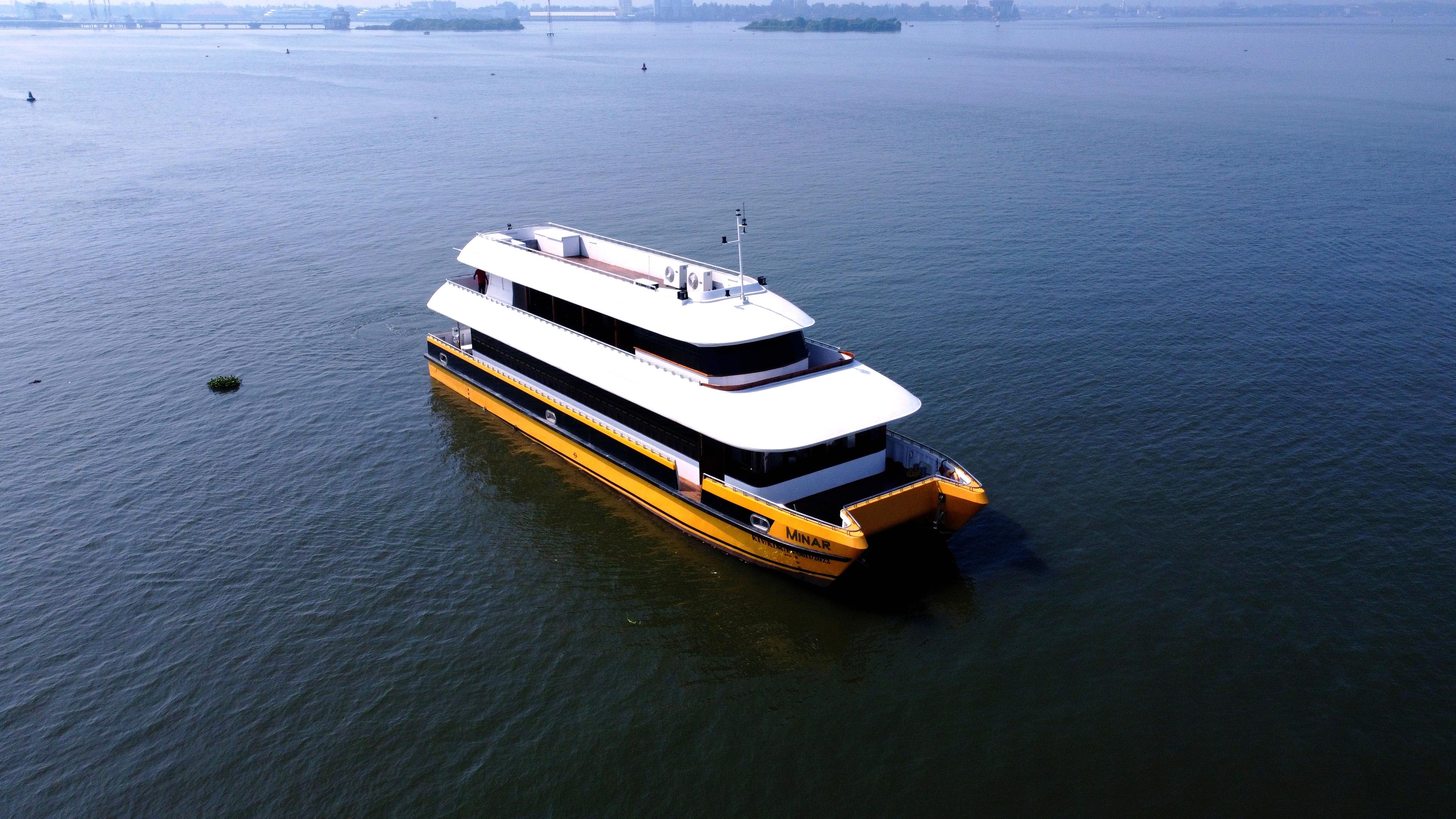 A yellow and white catamaran named Minar Cruise sailing on the calm waters of a bay with lush green mountains in the background in arabian sea, kochi, kerala, india.