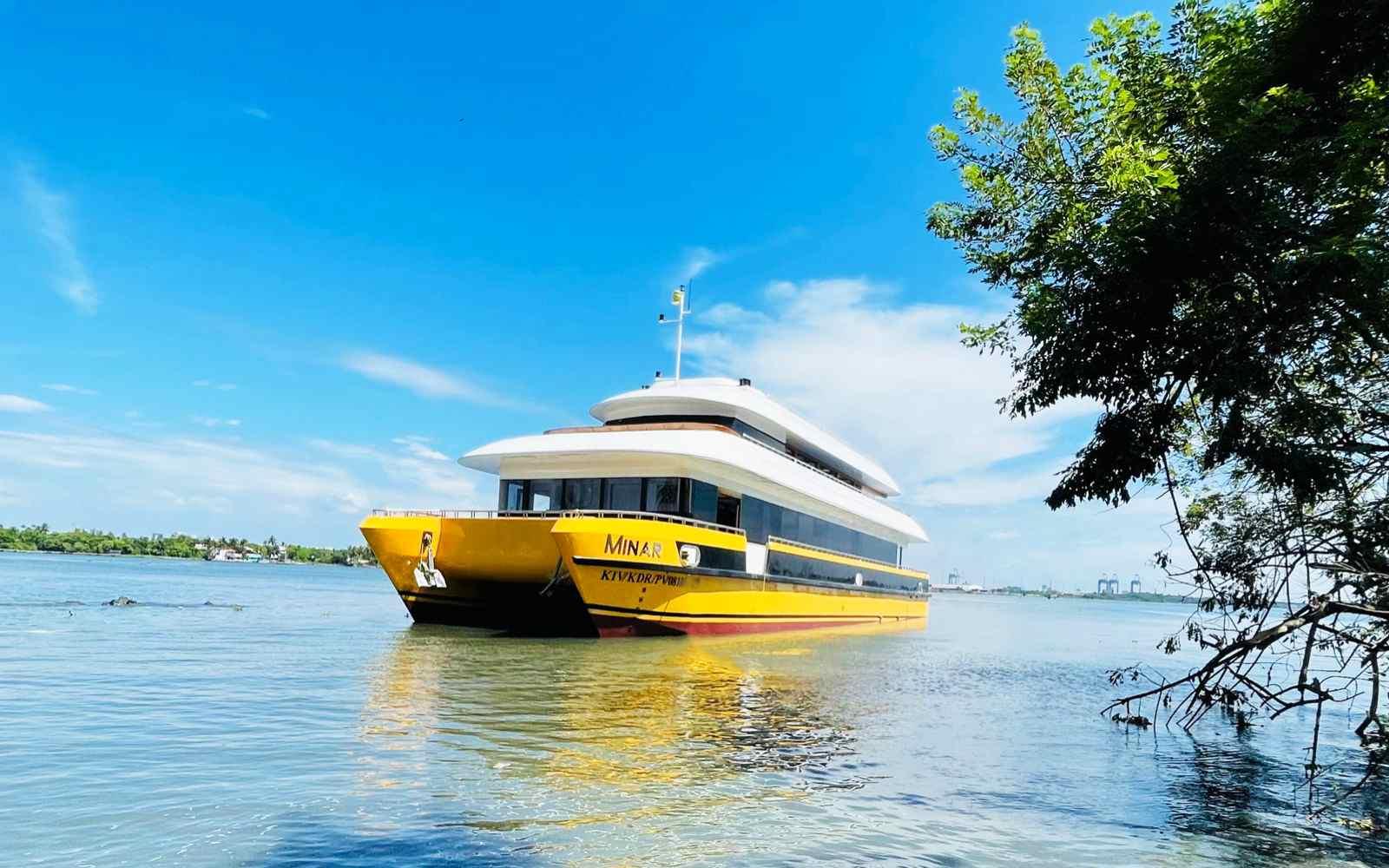 Frontview of A yellow and white catamaran named Minar Cruise sailing on the calm waters of a bay with lush green mountains in the background in arabian sea, kochi, kerala, india